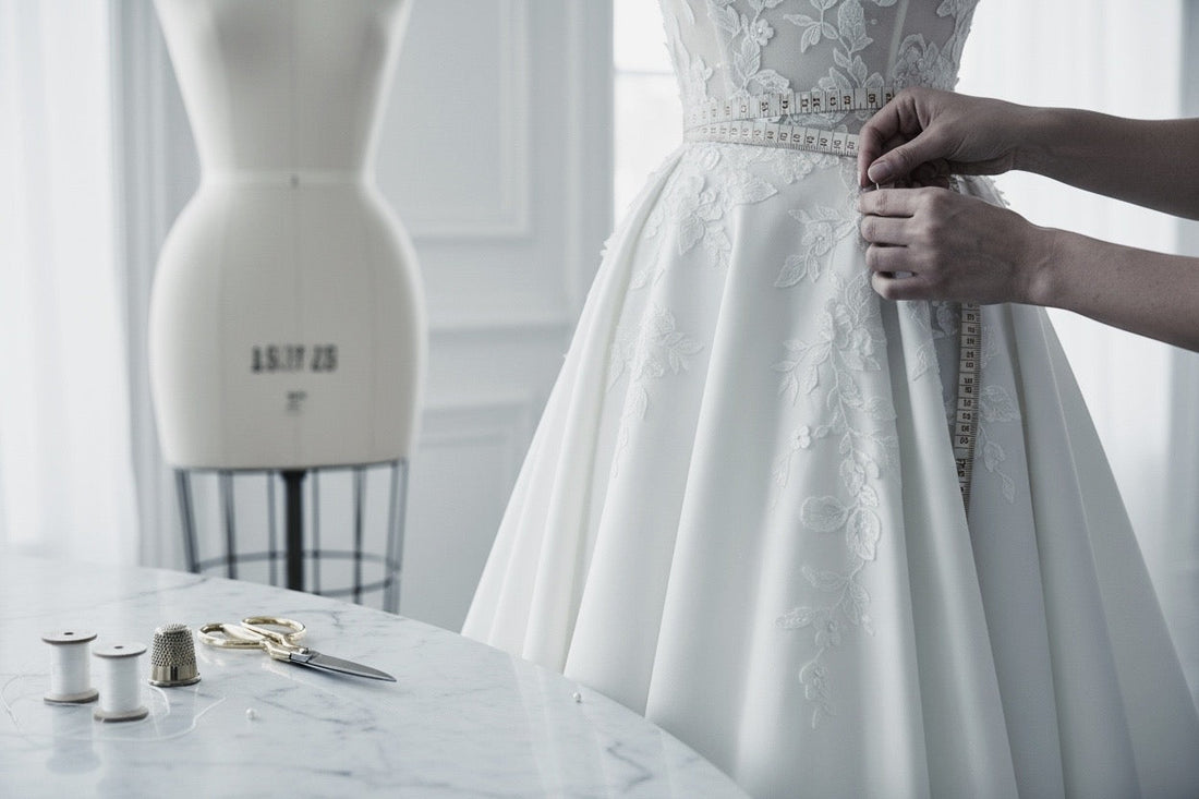 A close-up of a white lace wedding gown being adjusted by hands, with a dress form and sewing tools visible in a softly lit room.