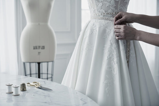 A close-up of a white lace wedding gown being adjusted by hands, with a dress form and sewing tools visible in a softly lit room.